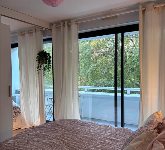 Chambre parentale élégante avec grandes fenêtres, rideaux toute hauteur et vue sur la végétation urbaine.